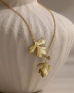 A gold pendant necklace featuring two textured metallic leaves hanging from a fine gold chain, displayed draped over a matte white ceramic vase.