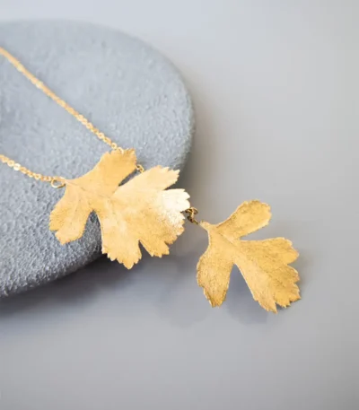 Close-up of a handmade necklace with two natural leaves preserved and plated in gold, attached to a fine gold chain, displayed on a round gray stone surface