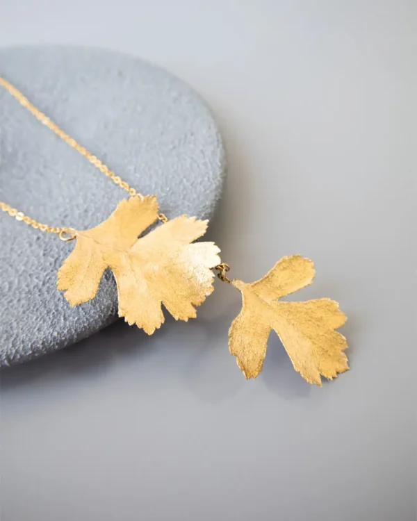 Close-up of a handmade necklace with two natural leaves preserved and plated in gold, attached to a fine gold chain, displayed on a round gray stone surface