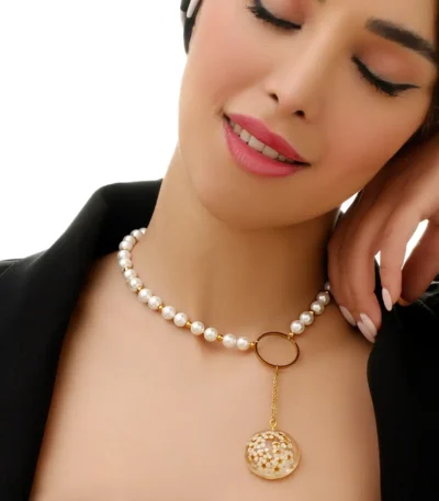 Close-up of a handmade pearl necklace with a golden chain and round pendant filled with tiny real dried flowers in resin, worn by a woman with closed eyes and natural makeup