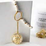 Handmade pearl necklace with golden ring detail and a round resin pendant filled with tiny dried white flowers, styled on a white block with a candle in the background
