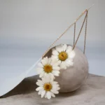 Artistic display of a handmade necklace featuring three real daisy flowers preserved in resin with golden centers, draped over a round stone on a minimalist background