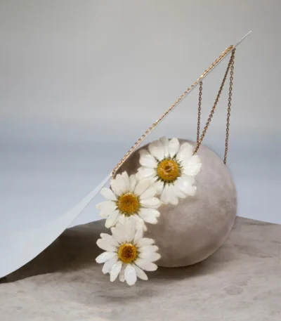 Artistic display of a handmade necklace featuring three real daisy flowers preserved in resin with golden centers, draped over a round stone on a minimalist background