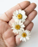 Close-up view of a handmade necklace with three real daisy flowers, preserved in resin, shown resting on a hand to highlight natural size and detail, with golden chain