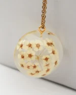Close-up view of a round resin pendant filled with delicate dried white flowers, hanging from a gold chain, handmade jewelry design