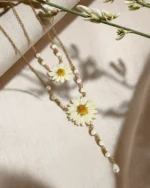 A delicate layered gold necklace featuring two real pressed daisy flowers and small white pearls, displayed on a soft beige fabric with natural light and floral shadows.