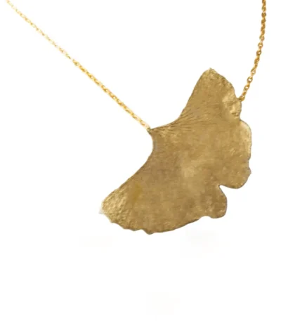 A close-up of a gold necklace featuring a single textured ginkgo leaf pendant hanging from a delicate gold chain on a white background