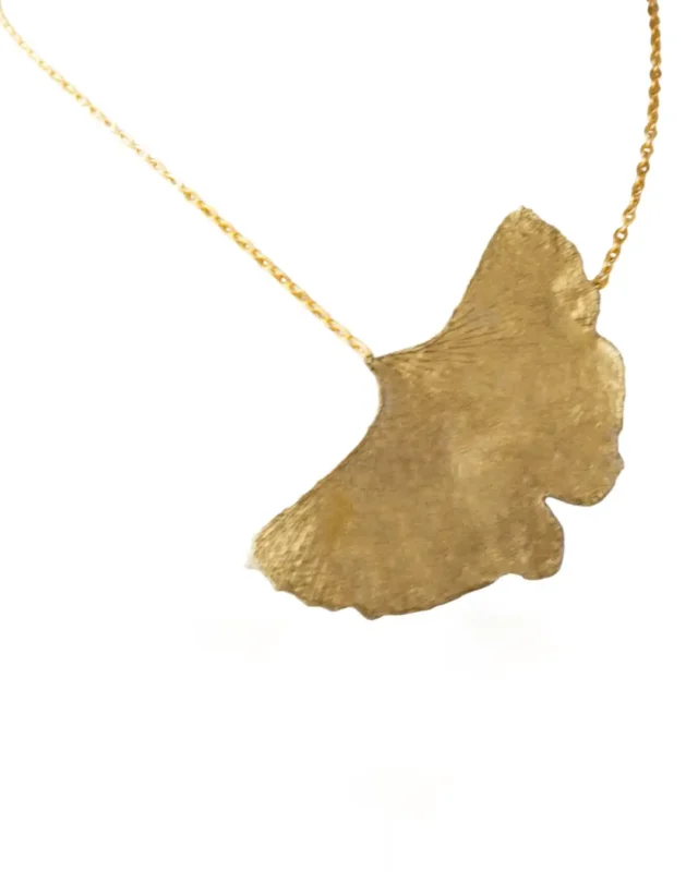 A close-up of a gold necklace featuring a single textured ginkgo leaf pendant hanging from a delicate gold chain on a white background