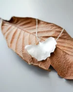 Handmade ginkgo leaf necklace with silver chain placed on dried brown leaf, nature-inspired jewelry design