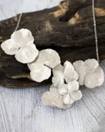 Handmade silver hydrangea flower necklace displayed against a piece of dark driftwood on a light textured surface.