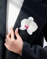 A white orchid brooch pinned to the lapel of a dark wool coat, worn by a person whose hand rests gently on the fabric