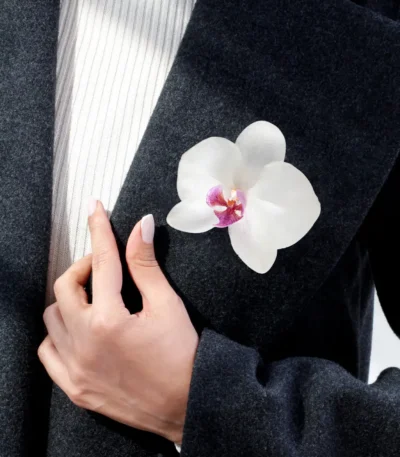 A white orchid brooch pinned to the lapel of a dark wool coat, worn by a person whose hand rests gently on the fabric