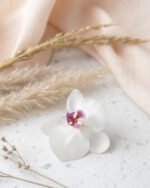Handmade white orchid flower with a soft purple center, crafted and preserved as jewelry, displayed on a light textured surface with beige fabric and dried grasses.